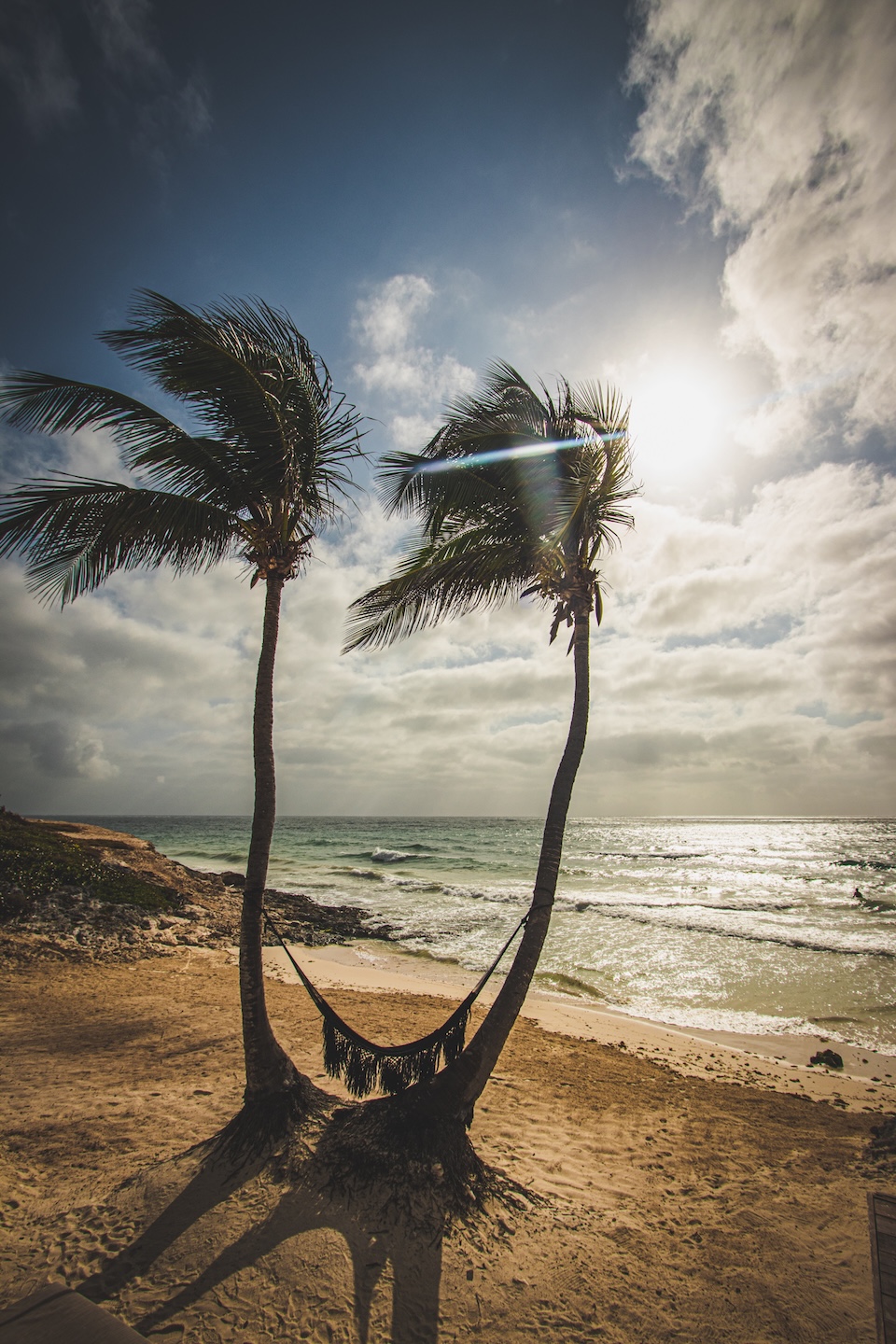 A hammock suspended between two palm trees on the beach in Tulum at sunrise, overlooking the calm sea and the golden morning light